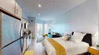 This single-floor studio is designed with an efficient open-concept layout that seamlessly integrates the sleeping, living, and kitchen areas into one cohesive space. Light wood-style flooring runs throughout, lending warmth and visual continuity across the entire unit.

The kitchen is well-appointed with stainless steel appliances, including a refrigerator, electric range, built-in microwave, and dishwasher. White cabinetry with sleek bar-pull hardware lines the upper and lower sections, complemented by dark granite-style countertops that add a modern contrast. An exposed cylindrical ductwork detail overhead introduces an industrial accent to the otherwise clean aesthetic.

The open living area accommodates a sofa and seating arrangement, flowing naturally into the sleeping space. Recessed lighting keeps the interior bright, while soft blue-gray walls create a calm, airy atmosphere. Sliding glass doors lead to a private balcony, allowing natural light to fill the space and providing outdoor access.

Toward the rear of the unit, a dedicated bathroom features a vanity with matching cabinetry and dark countertop, a large mirror, and wood-look tile flooring. A closet with mirrored sliding doors is also accessible near the sleeping area, offering practical storage within the compact footprint.