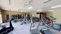 This fitness center is a comprehensive, single-floor facility designed to support a full range of workout routines. The main exercise area is generously sized, housing an array of cardio equipment including treadmills, ellipticals, and stationary and recumbent bikes, as well as multi-station strength training machines. The space features a distinctive carpet layout with red accent strips set against a neutral gray base, adding visual definition to the floor. Large wall-mounted mirrors extend across multiple sections of the room, enhancing the sense of space and allowing users to monitor their form during exercise. Multiple ceiling fans are distributed throughout, supplementing the fluorescent lighting and promoting air circulation during workouts. A wall-mounted television provides entertainment for cardio sessions. Natural light filters in through a series of arched windows along one wall, offering views to the outdoors and creating a bright, inviting atmosphere. A separate, open-plan area with the same red-accented carpet provides additional room for stretching or floor exercises. Motivational fitness posters and a wall clock complete the practical, purpose-driven décor. The textured ceiling and warm beige walls give the facility a clean, functional aesthetic throughout.