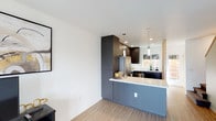Located at 2799 Adrian in San Diego, this two-story penthouse offers a thoughtfully designed living space with contemporary finishes and an efficient, modern aesthetic. The kitchen serves as a central highlight, featuring dark, flat-front cabinetry that extends to the ceiling for maximum storage, paired with a clean white countertop peninsula that doubles as a breakfast bar or casual dining surface. Pendant lighting hangs above the peninsula, providing focused task lighting while contributing to the modern ambiance. Stainless steel appliances, including a refrigerator and dishwasher, are seamlessly integrated into the cabinetry layout. A window above the sink allows natural light to filter into the space, brightening the work area. The flooring throughout is a warm-toned wood-look material that complements the cool gray and dark tones of the cabinetry. Crisp white walls keep the interior feeling open and airy despite the compact footprint. Recessed lighting supplements the pendant fixtures, ensuring even illumination throughout. The two-story layout adds a sense of vertical dimension, enhancing the overall spaciousness of this San Diego penthouse.