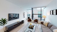 This well-appointed single-floor residence showcases a thoughtfully designed open-concept layout with a clean, contemporary aesthetic throughout. Light wood flooring flows seamlessly from room to room, complementing the crisp white walls and creating a warm, cohesive atmosphere.

The kitchen is a functional centerpiece, outfitted with gray flat-front cabinetry, stainless steel appliances including a gas range and full-size refrigerator, a white countertop, and a tiled backsplash. A peninsula with pendant lighting and bar seating doubles as a casual dining area, making the space ideal for both cooking and entertaining.

The living area is generously proportioned and bathed in natural light from large floor-to-ceiling windows that frame urban views of the surrounding cityscape. The open layout accommodates a comfortable seating arrangement with ample room to relax or host guests.

Two bedrooms offer restful retreats, each with light wood flooring and large windows that bring in natural light and city views. The primary bedroom features a striking teal upholstered headboard and stylish wall décor, while the second bedroom offers a similarly comfortable layout.

The bathroom is finished in an all-white palette, featuring a soaking tub with a tiled surround, a dual-sink vanity with white cabinetry, and a large mirror with overhead lighting, exuding a clean, spa-like quality.