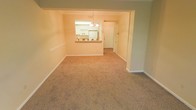 This single-floor residence offers a comfortable and functional layout designed for easy everyday living. The main living and dining area is finished with wall-to-wall carpeting in a warm, neutral tone, providing a soft and welcoming underfoot feel throughout the space. Walls are painted in a light, warm beige palette that lends a bright and airy ambiance to the interior.

A defining architectural feature is the pass-through window connecting the living and dining area to the kitchen, promoting an open and connected feel while maintaining a defined separation between spaces. The kitchen is finished with white cabinetry and includes a white refrigerator, creating a clean and classic look. A chandelier is suspended above the pass-through area, adding a decorative touch that enhances the dining experience.

The entry door is visible from the main living area, offering a clear and convenient sightline to the front of the home. White baseboards throughout provide a crisp contrast to the carpeted floors and painted walls. The overall layout feels cohesive and well-proportioned, making it a practical and inviting single-level living space suited for a variety of lifestyles.