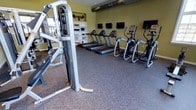 This dedicated fitness center offers a comprehensive workout environment designed to accommodate a range of exercise needs. The space is finished with durable rubber flooring, providing a safe and supportive surface for both cardio and strength training activities. A row of treadmills lines the main floor area, accompanied by elliptical cross-trainers, ensuring ample options for cardiovascular exercise. A multi-station strength training machine anchors one side of the room, providing versatility for resistance-based workouts. Wall-mounted flat-screen televisions are positioned throughout, allowing users to stay entertained during their sessions. Built-in open shelving along one wall offers organized storage for workout accessories and equipment. The walls are painted in a warm green tone, contributing to an energizing atmosphere, while exposed industrial-style ductwork runs along the ceiling, lending the space a modern, utilitarian aesthetic. Recessed lighting ensures the area is well-lit throughout. The overall layout is thoughtfully arranged to maximize usable floor space while maintaining clear pathways between equipment, creating a comfortable and functional environment for fitness enthusiasts of all levels.