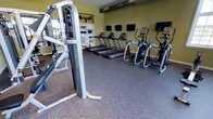 This dedicated fitness center offers a comprehensive workout environment designed to accommodate a range of exercise needs. The space is finished with durable rubber flooring, providing a safe and supportive surface for both cardio and strength training activities. A row of treadmills lines the main floor area, accompanied by elliptical cross-trainers, ensuring ample options for cardiovascular exercise. A multi-station strength training machine anchors one side of the room, providing versatility for resistance-based workouts. Wall-mounted flat-screen televisions are positioned throughout, allowing users to stay entertained during their sessions. Built-in open shelving along one wall offers organized storage for workout accessories and equipment. The walls are painted in a warm green tone, contributing to an energizing atmosphere, while exposed industrial-style ductwork runs along the ceiling, lending the space a modern, utilitarian aesthetic. Recessed lighting ensures the area is well-lit throughout. The overall layout is thoughtfully arranged to maximize usable floor space while maintaining clear pathways between equipment, creating a comfortable and functional environment for fitness enthusiasts of all levels.