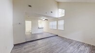 This single-floor unit is defined by its striking vaulted ceilings, which lend a sense of openness and volume to the main living area. Grey wood-style laminate flooring runs throughout the primary living spaces, complementing the neutral, cream-toned walls and creating a cohesive, modern aesthetic. Large windows fitted with vertical blinds allow natural light to fill the living and dining areas, enhancing the airy feel created by the elevated ceiling line. A ceiling fan with integrated light fixture serves the lower-ceilinged dining and kitchen zone, which transitions smoothly from the main living space.

The kitchen features white cabinetry, dark granite-style countertops, and a full suite of white appliances, including a refrigerator, dishwasher, and electric range. The bedroom areas include plush carpeted flooring and are equipped with bi-fold closet doors providing ample storage. One bedroom features dual closets with wire shelving. The bathroom includes a tub-shower combination, a vanity with dark countertop, and wood-style flooring. The overall layout is functional and well-organized, with a clean, neutral palette throughout.
