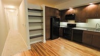 This single-floor residence offers a thoughtfully designed layout with a clean, modern aesthetic. The kitchen is a standout feature, showcasing rich dark espresso-toned cabinetry that extends to the upper walls, providing ample storage. Black appliances — including a full-size refrigerator, electric range, and built-in microwave — lend a sleek, cohesive look to the space. Light-toned countertops provide a subtle contrast against the dark cabinetry, while warm, natural hardwood flooring runs throughout, adding character and continuity. A built-in open shelving unit adjacent to the kitchen offers additional display and storage space, framed neatly between white trim columns. The warm yellow-toned walls complement the wood tones and create an inviting, comfortable atmosphere. The layout feels open and functional, with clear sightlines that enhance the sense of space. Overall, the residence balances practicality with style, featuring quality finishes and classic design elements that create a welcoming and well-organized living environment.