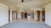 This single-story home showcases an open-concept layout that seamlessly connects the living area and kitchen into one cohesive space. The vaulted ceiling adds a sense of vertical volume, while a ceiling fan provides both function and a finishing touch overhead. Pendant lighting hangs above the kitchen zone, complementing the recessed lighting that illuminates the space evenly.

The kitchen is outfitted with light oak cabinetry, a central island for additional prep and storage space, and black appliances including a refrigerator, range, and dishwasher. The flooring transitions from neutral tile in the kitchen and dining zone to plush carpet in the living area, clearly defining each functional space while maintaining a unified, warm aesthetic.

Multiple interior doors are visible, suggesting easy access to additional rooms and storage areas throughout the home. The overall color palette relies on soft beiges and warm wood tones, creating a welcoming and practical environment. The layout feels spacious and well-organized, making it well-suited for everyday living and entertaining.