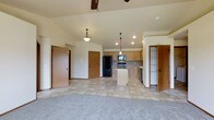 This single-story home showcases an open-concept layout that seamlessly connects the living area and kitchen into one cohesive space. The vaulted ceiling adds a sense of vertical volume, while a ceiling fan provides both function and a finishing touch overhead. Pendant lighting hangs above the kitchen zone, complementing the recessed lighting that illuminates the space evenly.

The kitchen is outfitted with light oak cabinetry, a central island for additional prep and storage space, and black appliances including a refrigerator, range, and dishwasher. The flooring transitions from neutral tile in the kitchen and dining zone to plush carpet in the living area, clearly defining each functional space while maintaining a unified, warm aesthetic.

Multiple interior doors are visible, suggesting easy access to additional rooms and storage areas throughout the home. The overall color palette relies on soft beiges and warm wood tones, creating a welcoming and practical environment. The layout feels spacious and well-organized, making it well-suited for everyday living and entertaining.