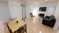 This single-floor residence offers a clean, functional layout designed for comfortable everyday living. The open-plan living and dining area is the heart of the space, unified by light hardwood flooring that extends throughout, lending a warm and airy feel to the interior.

The dining area accommodates a wooden table with seating for multiple guests, while a small decorative vase adds a touch of warmth. The space flows seamlessly into the living area, which features a wall-mounted flat-screen television, a fireplace that serves as a natural focal point, and a glass-top coffee table surrounded by leather seating. A sleek accent chair completes the arrangement, providing additional seating options.

The crisp white walls and recessed lighting contribute to a bright, neutral ambiance throughout. A hallway visible from the main living area leads to additional rooms, suggesting a thoughtful and practical floor plan. The overall design is minimalist yet inviting, with clean lines and an uncluttered aesthetic that makes the space feel open and well-proportioned. The combination of the fireplace, coordinated furnishings, and consistent flooring creates a cohesive and welcoming environment suitable for both relaxing and entertaining.