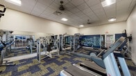 This fitness center is a fully outfitted exercise facility designed to support a complete workout regimen. The space is anchored by a bold, navy blue accent wall adorned with a health and wellness-themed word mural featuring terms like "health," "fitness," "lifestyle," and "energy," creating an energizing and motivational atmosphere throughout.

The cardio zone includes multiple treadmills and elliptical trainers, all positioned to allow comfortable use with ample space between machines. The strength training area is equally well-appointed, featuring a multi-station cable machine, a functional trainer, a variety of selectorized resistance machines, a dumbbell rack, weight benches, and a dedicated free weights section. Full-length mirrors line portions of the walls, enhancing the sense of space and allowing users to monitor their form during workouts.

Flooring consists of durable patterned modular carpet tiles in blue and tan tones, providing cushioning and visual interest. The drop ceiling features recessed lighting and ceiling fans to ensure a well-lit and ventilated environment. Arched windows along one wall invite natural light and views of surrounding greenery. A flat-screen television is wall-mounted for entertainment. Glass sliding doors connect the fitness center directly to an adjacent lounge and common area, offering seamless access to additional amenities.
