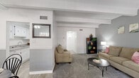 This thoughtfully designed single-floor residence offers a comfortable and functional layout across its key living spaces. The combined living and dining area forms the heart of the unit, featuring soft carpeted flooring, exposed beam ceiling details, and neutral gray walls that create a calm, cohesive atmosphere. The living room benefits from a large window that invites natural light and outdoor views, while decorative beams add architectural character overhead. Adjacent to the living room, the dining area is finished with light hardwood-style flooring, a ceiling fan, wainscoting along the lower walls, and a sliding glass door that further brightens the space with natural light. The galley-style kitchen is efficiently designed with ample white cabinetry, granite-style countertops, a double sink, and white appliances, all set atop the same light hardwood-style flooring. The spacious bedroom is a standout feature, showcasing a striking white-painted brick accent wall, exposed ceiling beams, plush carpeting, and a large window. A hallway connects the bedroom to the full bathroom, which features updated countertops and bright lighting, alongside a separate closet with double doors for generous storage.