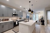 This single-floor residence offers a thoughtfully designed open-concept layout that seamlessly connects the kitchen, dining, and living areas. The kitchen is a standout feature, showcasing gray shaker-style cabinetry extending to the ceiling, white quartz countertops, a classic subway tile backsplash, and a full suite of stainless steel appliances. A central island with an undermount sink and striking black pendant lights above serves as both a functional workspace and a natural gathering point. Light wood-look flooring runs throughout the main living areas, enhancing the bright, airy atmosphere.

The living room is welcoming and well-lit, benefiting from natural light streaming through French doors that open to a private balcony. A ceiling fan overhead and neutral-toned walls contribute to a calm, comfortable ambiance.

The bedroom continues the refined aesthetic with plush carpeting, a ceiling fan, large windows, and a bold navy accent wall that adds depth and character to the space.

The bathroom is elegantly appointed with gray shaker vanity cabinetry, a white quartz countertop, matte black hardware, a large framed mirror, and a glass-enclosed walk-in shower with large-format tile. Light wood-look flooring ties the space together cohesively.
