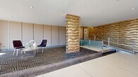 Located at 243 S Harrison St in East Orange, NJ, this building lobby is a well-appointed, single-floor entry space that makes a strong architectural first impression. The centerpiece of the lobby is a dramatic floor-to-ceiling column clad in a richly textured, wave-patterned gold and bronze material that draws the eye immediately upon entry. Matching accent walls with the same undulating texture reinforce the design language throughout the space.

The flooring transitions between large-format terrazzo tiles in the entry zone and a bold, geometric-patterned carpet on the raised interior platform, adding visual depth and defining distinct areas within the lobby. Recessed ceiling lighting provides even, modern illumination throughout.

Floor-to-ceiling glass at the main entrance floods the space with natural light and offers views of the surrounding streetscape. A sleek seating area with upholstered chairs in muted tones and a marble-top table offers a comfortable waiting area for residents and guests. 

A glass-panel staircase with stainless steel railings leads to upper floors, adding a contemporary, refined touch. The overall ambiance is modern and polished, blending decorative flair with functional design to create a sophisticated and welcoming arrival experience.
