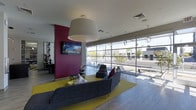 This single-floor space features a contemporary lounge area designed with a bold and energetic aesthetic. A striking magenta accent wall serves as a focal point, anchoring a wall-mounted flat-screen television and adding a vibrant contrast to the neutral grey ceiling and light hardwood-style flooring throughout.

The seating area is centered around a yellow area rug, which grounds a grouping of modern grey sofas and accent chairs, creating an inviting conversation zone. Overhead, a large drum pendant light with a white shade commands attention, complemented by smaller globe pendant fixtures that contribute to a layered lighting scheme.

An expansive floor-to-ceiling glass window wall runs along one side of the space, flooding the interior with abundant natural light and offering open views of the surrounding streetscape. This transparent boundary creates a seamless visual connection between the interior and the exterior environment.

Additional seating arrangements are visible near the windows, providing flexible options for occupants. The overall design balances bold color choices with clean, modern lines, resulting in a dynamic yet functional communal space suited for lounging, socializing, or casual gatherings.