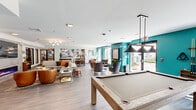 This single-floor amenity lounge is a thoughtfully designed communal space that blends social entertainment with comfortable relaxation. The open-plan layout is anchored by a full-size pool table positioned beneath a sleek pendant light fixture, serving as the centerpiece of the room. Bold teal accent walls provide a vibrant contrast to the light-toned luxury vinyl plank flooring and crisp white ceilings, creating a modern and energetic atmosphere throughout.

Multiple distinct seating areas are arranged across the space, each offering its own character — from plush sofas and round accent chairs in warm cognac leather to clusters of upholstered armchairs ideal for conversation. A built-in kitchenette is outfitted with a refrigerator, microwave, and counter space, backed by a striking abstract mural that adds an artistic focal point to the area.

A separate lounge nook features a linear gas fireplace set into a tile surround with a wall-mounted television above, flanked by additional seating for a more intimate gathering experience. Large windows and glass doors along the teal-accented wall flood the interior with natural light and provide direct views and access to an outdoor patio area. The overall ambiance is lively yet inviting, making it an ideal multifunctional space for both recreation and community connection.