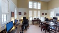 Located at 900 Beverly Hills Rd in Hattiesburg, Mississippi, this single-floor business center offers a welcoming and functional environment designed to meet residents' productivity needs. The space is immediately distinguished by its dramatically high ceilings, which lend an open, airy feel throughout the room. Large, multi-pane windows line the walls and rise nearly to the ceiling, flooding the interior with generous natural light while Roman shades provide adjustable privacy and light control. The walls are finished in a warm cream tone with classic beadboard wainscoting, contributing to a clean and inviting aesthetic. A row of computer workstations with full-size monitors and keyboards lines one wall, providing ample workspace for individual use. On the opposite side, a dedicated coffee bar station offers a convenient amenity for visitors. A seating area with dark wood chairs and a round table provides a comfortable spot for casual meetings or relaxed work sessions. Decorative wall art with a tech-inspired theme adds personality to the space. The light-toned carpeting ties the room together, creating a cohesive and comfortable atmosphere that balances professionalism with a warm, community-oriented charm.