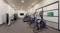 This fitness center offers a comprehensive and thoughtfully arranged workout environment designed to support a wide range of exercise routines. The space is laid out on a single level, with recessed lighting illuminating the clean, white walls and durable rubberized flooring throughout. A row of cardio machines lines one side of the room, including multiple treadmills equipped with individual screens, elliptical trainers, and a stair climber, providing ample options for cardiovascular training. A wall-mounted flat-screen television is positioned for easy viewing during workouts. On the strength-training side, a multi-functional cable machine serves as a centerpiece, outfitted with numerous attachments to accommodate a variety of exercises. A well-stocked dumbbell rack with a full range of weights sits nearby, catering to both beginner and advanced lifters. Full-length mirrors along one wall visually expand the space and allow users to monitor their form. Black-framed glass partitions add a modern architectural touch, while a window introduces natural light. The overall ambiance is clean, modern, and functional, creating an inviting environment for focused, effective training.