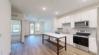 This single-floor unit presents a clean, contemporary design with a well-considered layout visible across its interconnected spaces. The kitchen serves as a central highlight, featuring white shaker-style cabinetry extending to the ceiling, a grey tile backsplash, stainless steel appliances including a refrigerator, range, microwave, and dishwasher, and stone-look countertops. A freestanding island with a marble-style white surface and dark wood frame provides additional workspace and casual dining potential. Wood-look flooring runs throughout the main living areas, lending warmth and continuity to the open-concept space.

The living area adjoins the kitchen seamlessly, with recessed lighting, a ceiling fan, and a glass door opening to a private balcony that brings in natural light and outdoor views. Multiple additional rooms feature plush neutral carpeting, ceiling fans, and windows fitted with horizontal blinds, offering comfortable, well-lit spaces suited for various uses. One room includes its own door to the balcony, further enhancing connectivity to the outdoors.

The bathroom is finished with white cabinetry, stone-look countertops, a large framed mirror, and a marble-style tiled shower surround, maintaining the unit's consistent modern aesthetic throughout. Overall, the space conveys a bright, move-in-ready atmosphere with cohesive finishes.