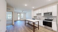 This single-floor unit presents a clean, contemporary design with a well-considered layout visible across its interconnected spaces. The kitchen serves as a central highlight, featuring white shaker-style cabinetry extending to the ceiling, a grey tile backsplash, stainless steel appliances including a refrigerator, range, microwave, and dishwasher, and stone-look countertops. A freestanding island with a marble-style white surface and dark wood frame provides additional workspace and casual dining potential. Wood-look flooring runs throughout the main living areas, lending warmth and continuity to the open-concept space.

The living area adjoins the kitchen seamlessly, with recessed lighting, a ceiling fan, and a glass door opening to a private balcony that brings in natural light and outdoor views. Multiple additional rooms feature plush neutral carpeting, ceiling fans, and windows fitted with horizontal blinds, offering comfortable, well-lit spaces suited for various uses. One room includes its own door to the balcony, further enhancing connectivity to the outdoors.

The bathroom is finished with white cabinetry, stone-look countertops, a large framed mirror, and a marble-style tiled shower surround, maintaining the unit's consistent modern aesthetic throughout. Overall, the space conveys a bright, move-in-ready atmosphere with cohesive finishes.