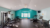 Located at 300 Caughman Farm Ln in Lexington, South Carolina, this single-floor fitness center offers a well-appointed and generously proportioned workout facility designed to support a comprehensive exercise routine. The space is immediately distinguished by its vaulted ceilings, which create an open and airy atmosphere, complemented by recessed lighting and a ceiling fan that ensure both adequate illumination and comfortable airflow throughout workouts. Bold teal accent walls add a vibrant, energizing character to the interior, contrasting cleanly with the white ceiling and trim. Durable rubber flooring covers the entire workout area, providing both safety and practicality underfoot. The facility is equipped with a variety of cardio machines, including treadmills, ellipticals, and a stationary cycle, as well as strength-training equipment and a dumbbell rack catering to a range of fitness needs. Wall-mounted televisions are positioned throughout the space for entertainment during exercise. A bay window along one wall allows natural light to filter into the room, brightening the space and offering views of the surrounding exterior. Additional doors provide access to adjoining areas, and a whiteboard is available for workout tracking or announcements. The overall ambiance is functional, bright, and motivating.