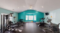 Located at 300 Caughman Farm Ln in Lexington, South Carolina, this single-floor fitness center offers a well-appointed and generously proportioned workout facility designed to support a comprehensive exercise routine. The space is immediately distinguished by its vaulted ceilings, which create an open and airy atmosphere, complemented by recessed lighting and a ceiling fan that ensure both adequate illumination and comfortable airflow throughout workouts. Bold teal accent walls add a vibrant, energizing character to the interior, contrasting cleanly with the white ceiling and trim. Durable rubber flooring covers the entire workout area, providing both safety and practicality underfoot. The facility is equipped with a variety of cardio machines, including treadmills, ellipticals, and a stationary cycle, as well as strength-training equipment and a dumbbell rack catering to a range of fitness needs. Wall-mounted televisions are positioned throughout the space for entertainment during exercise. A bay window along one wall allows natural light to filter into the room, brightening the space and offering views of the surrounding exterior. Additional doors provide access to adjoining areas, and a whiteboard is available for workout tracking or announcements. The overall ambiance is functional, bright, and motivating.
