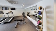 This fitness center offers a comprehensive workout environment designed to support a wide range of exercise routines. The space is laid out across a single floor and features durable, speckled rubber flooring throughout, providing both safety and comfort during workouts. A large floor-to-ceiling mirror runs along one wall, visually expanding the space and allowing users to monitor their form during exercise.

The cardio section is equipped with multiple Spirit treadmills and an elliptical trainer, positioned near large windows fitted with horizontal blinds that allow natural light to filter in. Mounted flat-screen televisions are positioned on the wall above the cardio machines, offering entertainment during workouts. A stationary bike and additional cardio equipment round out the aerobic offerings.

The strength training area includes a multi-position adjustable workout bench, and a well-stocked rack of dumbbells in graduated weights, suitable for a variety of resistance exercises. A dedicated medicine ball rack holds an assortment of weighted medicine balls in multiple sizes, supporting functional and core training needs.

The center features a textured ceiling with recessed lighting and track lighting that keep the space bright and functional. Posted fitness center rules on the wall reflect an organized, community-oriented facility. The overall ambiance is clean, practical, and purposefully arranged to maximize usability.