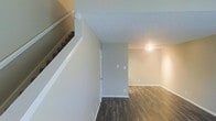 This two-story space offers a straightforward and functional layout designed with simplicity and practicality in mind. The main level features rich, dark hardwood-style flooring that contrasts beautifully with the light neutral gray walls, creating a warm yet contemporary aesthetic throughout. White baseboards and trim provide a crisp finishing touch to the clean interior palette.

A staircase with a white painted handrail connects the two floors, positioned to allow an open sightline across the main level. The textured ceiling adds subtle visual depth while maintaining a modest, understated character throughout the space.

The open living area on the main floor benefits from overhead lighting and is anchored by wide, open walls that offer flexible furniture arrangement. A doorway leads to an additional room or exit, adding to the practical flow of the floor plan.

The neutral color scheme of warm grays and whites throughout provides a versatile backdrop suitable for a variety of interior styles. The combination of durable flooring, clean walls, and a well-considered two-floor layout makes this space both functional and inviting. Overall, the interior presents a move-in ready environment with a modern, no-frills design sensibility ideal for everyday living.