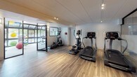 This fitness center offers a clean, modern environment designed to support a well-rounded workout routine. The space is finished with warm wood-look flooring and white walls, complemented by recessed track lighting that keeps the area bright and functional throughout.

The cardio zone is equipped with multiple treadmills and elliptical trainers arranged along one side of the room, providing ample space between machines for comfortable use. A multi-functional cable trainer station anchors another area of the floor, suitable for a wide range of strength and resistance exercises. A flat weight bench accompanies this zone, adding versatility for free-weight training.

A glass-partitioned section of the fitness center creates a dedicated open-floor studio space, ideal for stretching, yoga, or bodyweight training. This area benefits from large windows that invite natural light and views of the surrounding greenery, contributing to an airy, welcoming atmosphere.

Safety is addressed with a clearly mounted AED defibrillator on the wall. The space also includes a private door leading to an additional room or utility area. Overall, the fitness center balances a contemporary aesthetic with practical, well-organized equipment placement to serve a variety of fitness needs.