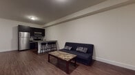 This well-appointed single-floor unit offers a thoughtfully designed open-concept layout that seamlessly connects the kitchen and living area. Rich hardwood-style flooring runs throughout the main living space, creating a warm and cohesive aesthetic. The kitchen is outfitted with dark espresso cabinetry, granite countertops, stainless steel appliances including a refrigerator, electric range, and over-the-range microwave, and a stylish mosaic tile backsplash. A peninsula with bar seating provides a casual dining option and further defines the kitchen zone within the open floor plan. The living area offers a comfortable, versatile space with clean white walls and ample room for various furniture arrangements. The bedroom is carpeted for added comfort and features windows with blinds that allow natural light to filter in, creating a bright and airy atmosphere. The bathroom is finished with large-format neutral tiles and features a spacious glass-enclosed walk-in shower with a mosaic tile floor, complemented by a wall-mounted vanity. The unit's overall design balances modern finishes with functional living spaces, making it a practical and stylish home.