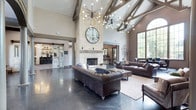 This clubhouse is designed to impress from the moment you enter, anchored by soaring vaulted ceilings accented with exposed wooden beams that lend a rustic yet refined character to the space. The dramatic ceiling height is further highlighted by two striking sputnik-style chandeliers, whose starburst silhouettes add a modern, industrial edge to the otherwise classic interior.

At the heart of the main lounge area stands a floor-to-ceiling stone fireplace, serving as the room's undeniable focal point. A large decorative clock mounted above the mantel reinforces the grandeur of the vertical space. Rich, dark polished concrete flooring runs throughout, reflecting light and contributing to the upscale ambiance.

Tufted leather sofas and Chesterfield-style seating are arranged to encourage conversation, complemented by area rugs that define the gathering zones. Large arched windows along one wall allow generous natural light to filter in, offering views of the surrounding greenery outside.

Toward the back, a barn-style door leads to an additional room, hinting at further amenities within the clubhouse. The overall design seamlessly blends rustic architectural elements with contemporary fixtures, creating a welcoming and stylish environment suited for both socializing and relaxation.