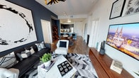This contemporary single-floor residence showcases a thoughtfully designed open-concept layout that connects the living, dining, and kitchen areas into one cohesive, flowing space. The living area is anchored by rich hardwood-style flooring and a striking deep navy accent wall that adds depth and sophistication to the room. A geometric chevron-patterned area rug grounds the seating arrangement, which features a sleek black sofa and a crisp white accent chair, complemented by a marble-top coffee table. An artistic sputnik-style chandelier overhead adds a bold, decorative focal point. The kitchen is sleek and modern, equipped with stainless steel appliances, dark cabinetry, and a vivid teal-blue island that serves as both a functional workspace and a visual centerpiece. The dining area is nestled between the living space and kitchen, furnished with contemporary chairs that echo the room's modern aesthetic. Throughout the space, track lighting ensures versatile illumination across all areas. The overall ambiance is dynamic and curated, blending bold color choices with clean architectural lines and high-end finishes to create a space that feels both stylish and functional.