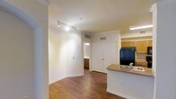 This single-floor space offers a clean, open-concept layout that connects the kitchen and living areas in a practical and comfortable arrangement. Rich hardwood flooring flows throughout, adding warmth and a sense of continuity across the space. The kitchen is positioned along one side and features honey-toned wood cabinetry, a black refrigerator, and a countertop with a built-in sink that extends into a peninsula, providing both workspace and a natural divider between the kitchen and living area. Track lighting is mounted on the ceiling of the main living space, offering adjustable, directed illumination across the room. The walls are finished in a neutral cream tone, complemented by white crown molding and baseboards that add a polished, classic touch throughout. A set of double doors with raised-panel detailing provides access to a closet, offering generous storage. A hallway visible in the background leads to what appears to be a bathroom, with warm wood cabinetry visible in the distance. Ceiling-mounted HVAC vents suggest a central climate control system. Overall, the space presents a bright, tidy, and functional interior with a neutral palette well-suited for a variety of furnishing styles.