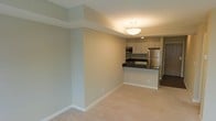 This single-floor space offers a clean, open-concept design that connects the living area and kitchen in a cohesive and functional layout. The living area is finished with plush neutral-toned carpeting and smooth walls painted in a soft beige palette, lending the space a calm and inviting ambiance. A structural soffit detail adds architectural character to the ceiling, while a central overhead light fixture provides warm, even illumination throughout.

The kitchen is thoughtfully designed with a raised breakfast bar featuring a dark countertop that serves as both a workspace and a casual dining surface. White cabinetry lines the upper walls, offering generous storage, while stainless steel appliances — including a refrigerator and built-in oven — bring a modern, polished aesthetic. The contrasting dark countertops against the white cabinetry create a striking visual detail. A door visible beyond the kitchen hints at additional rooms or storage areas within the unit.

The overall design emphasizes practicality and style, with consistent neutral tones throughout that create a bright and versatile backdrop suitable for a variety of interior arrangements. The open flow between the kitchen and living space makes the unit feel expansive and well-organized.