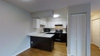 This single-floor space offers a clean, modern aesthetic anchored by a thoughtfully designed kitchen. The kitchen features crisp white upper cabinetry that contrasts elegantly with dark lower cabinets, creating a bold two-tone look. A marble-style countertop extends into a peninsula-style breakfast bar, providing additional prep space and a casual dining area. The kitchen is equipped with black appliances, including a refrigerator, electric range, microwave, and a stainless steel sink, all set beneath a textured ceiling with flush-mount lighting. Warm hardwood-style flooring flows throughout, adding a sense of cohesion and warmth to the space. A louvered closet door is visible near the kitchen, suggesting convenient built-in storage. The neutral gray wall tones complement the overall color palette, keeping the space feeling bright and contemporary. The layout is efficient and functional, making excellent use of the available footprint. Overall, the space presents a refreshed, move-in-ready interior with modern finishes that balance style and practicality throughout.