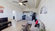 This single-floor unit offers a well-organized layout that maximizes every square foot of its cozy footprint. The open-concept main area seamlessly integrates the living room and kitchen, creating a functional and welcoming space. Light wood-look flooring runs throughout, complemented by crisp white walls and exposed ceiling beams that add architectural character. Ceiling fans are installed in multiple rooms, promoting airflow and comfort throughout.

The kitchen is equipped with gray cabinetry, a black gas range, a full-size refrigerator, a microwave, and a sink — providing all essential cooking amenities in a compact arrangement. The living area accommodates a sectional sofa, a coffee table, a dining set for two near a window, and a television setup, making efficient use of the shared space. Natural light filters in through windows dressed with gray curtain panels.

The bedroom is a comfortable retreat featuring exposed painted brick walls, a ceiling fan with light, a window with an individual climate control unit, and direct access to a walk-in closet. The bathroom includes a vanity with a black-finish faucet, a large mirror, black hardware accents, and a shower with a tiled surround — finished with light flooring that ties the space together cohesively.