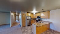 Located at 2875 36th Ave S in Grand Forks, North Dakota, this single-floor unit offers a thoughtfully designed layout with a range of functional features throughout. The kitchen serves as a central highlight, showcasing warm oak cabinetry, granite-style countertops, and a full suite of stainless steel appliances including a refrigerator, electric range, and over-the-range microwave. A peninsula with a built-in sink and pendant lighting overhead adds both style and practicality, while tile flooring ties the space together and flows seamlessly into the adjoining living area. A long hallway connects the main living spaces to the private areas of the unit, where multiple carpeted bedrooms provide comfortable retreats. Each bedroom features neutral wall tones, wood-trimmed windows with blinds, and views of the surrounding neighborhood. The bathroom is well-appointed with oak vanity cabinetry, granite-style countertops, a tall linen tower, and a walk-in shower enclosed by sliding glass doors, finished with tile flooring for easy maintenance. A dedicated in-unit laundry area accommodates a full-size washer and dryer side by side, complete with overhead shelving for added storage. The overall aesthetic is clean and consistent, with warm wood accents and neutral tones creating a cohesive and livable environment throughout.