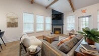 Located at 330 Cunningham Lane in Clarksville, Tennessee, this single-floor space offers a thoughtfully designed layout that blends comfort and functionality. The open-concept kitchen and living room serve as the heart of the space, creating a seamless flow between cooking and gathering areas. The kitchen is outfitted with crisp white shaker cabinets, dark stone countertops, a decorative tile backsplash, and black appliances, including a side-by-side refrigerator and dishwasher, all set atop warm hardwood-style flooring. Natural light filters in through multiple windows dressed with white blinds, brightening the space throughout the day. The living room is a standout, featuring vaulted ceilings accented by rustic exposed wood beams that add warmth and architectural character. A striking dark shiplap fireplace surround with an electric insert serves as the focal point, flanked by large windows that invite ample natural light. The comfortable seating arrangement and area rug create an inviting atmosphere well-suited for relaxation. A dedicated exercise room provides a private space for fitness activities, while a connecting hallway ties the layout together efficiently. Overall, the space strikes a balance between rustic charm and modern finishes.