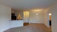 This single-floor space showcases an open-concept layout that connects the kitchen and living area in a cohesive, functional design. The living area is finished with plush carpeting, offering a comfortable and inviting atmosphere, while the kitchen and entryway transition to smooth hardwood-style flooring, creating a natural visual distinction between the zones.

The kitchen features dark cabinetry, a granite-topped breakfast bar that serves as both a workspace and casual dining counter, and a track lighting system that provides focused illumination over the cooking area. A pendant light with a classic bowl-shaped fixture hangs elegantly in the transitional space, adding a decorative touch while enhancing overall brightness.

The entryway is clearly defined, with a paneled white door that anchors the space and provides a welcoming focal point. Neutral white walls throughout contribute to a clean, airy feel, allowing the warm overhead lighting to set a cozy tone. The open flow between areas makes the space feel generously proportioned, well-suited for both everyday living and entertaining.
