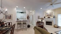 This single-floor residence offers an open and functional layout that seamlessly connects the kitchen and living areas. The kitchen features white cabinetry with raised-panel detailing, a built-in microwave, and a white refrigerator. A peninsula with a white countertop extends into the living space, creating a casual breakfast bar equipped with two elegant wrought-iron stools, making it an ideal spot for informal dining or conversation.

The living area is warm and inviting, anchored by a classic fireplace with a white mantel that serves as a natural focal point. A decorative console table positioned nearby holds a lamp and small accents, adding a refined touch to the space. Framed artwork on the walls and recessed lighting throughout contribute to the bright, welcoming atmosphere.

Neutral carpet and light-colored walls are consistent throughout, lending a cohesive and airy feel to the interior. A hallway visible from the main living area suggests additional rooms beyond. The overall design balances practicality with comfort, making it a well-appointed and livable single-floor home.