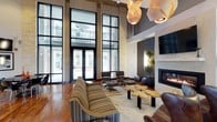 This single-floor space makes an immediate impression with its dramatic, soaring ceiling height and expansive floor-to-ceiling windows framed in dark steel. The oversized glass panels flood the interior with natural light while offering open views to an outdoor courtyard, creating a seamless connection between inside and out. Warm hardwood flooring anchors the space and contrasts beautifully with the natural stone and textured wall elements throughout.

The lounge area is thoughtfully appointed with a curated mix of furnishings, including a boldly striped sofa, classic leather lounge chairs with ottomans, and organically shaped wood coffee tables that add an artistic, nature-inspired touch. A built-in fireplace set within a textured stone surround serves as a focal point on one wall, flanked by live greenery and topped with a wall-mounted display screen, balancing warmth with modern functionality.

Overhead, sculptural pendant lighting in warm tones adds an artistic dimension to the space, complementing the industrial-chic aesthetic of the steel window frames and exposed structural elements. The overall ambiance is one of refined comfort — a space that is both visually striking and inviting, ideal for gathering, relaxing, or socializing in a stylish, well-designed environment.