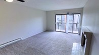 This single-floor space offers a clean, functional layout designed for comfortable living. The main living area is generously sized, finished with wall-to-wall carpeting that adds warmth and softness underfoot. Neutral-toned walls provide a versatile backdrop, making the space easy to personalize. A textured ceiling adds subtle visual interest overhead, while a ceiling fan ensures year-round comfort.

The standout feature of the living area is the large sliding glass door that floods the room with natural light and provides direct access to a private balcony. The balcony offers open-air views of the surrounding building exterior, creating a pleasant outdoor extension of the living space. Sheer vertical blinds frame the glass door, offering flexible light control throughout the day.

The transition from carpeted living area to hard flooring near the entryway provides a practical and well-defined boundary between spaces. The overall ambiance is bright, airy, and move-in ready, with a straightforward layout that maximizes usable space. Clean lines, minimal architectural detailing, and ample natural light give this space a simple yet inviting character well-suited for a variety of living arrangements.