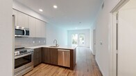 Located on Westwood Boulevard in Los Angeles, this modern single-floor unit offers a clean, contemporary design throughout. The open-concept living and kitchen area serves as the heart of the space, where warm hardwood-style flooring flows seamlessly from room to room. The kitchen is thoughtfully designed with two-tone cabinetry—white upper cabinets and rich wood-grain lower cabinets—paired with white quartz countertops, a tile backsplash, and a full suite of stainless steel appliances including a gas range, microwave, dishwasher, and refrigerator. A peninsula with an undermount sink adds both functionality and definition between the kitchen and the open living area. The living space is bright and airy, with recessed lighting and direct access to a private balcony through full-height glass doors that invite natural light and views of lush surrounding greenery. A dedicated bedroom area features its own balcony access and a spacious walk-in closet with built-in shelving. The bathroom is finished with large-format neutral floor tiles, white subway wall tiles, a soaking tub with shower, matte black fixtures, and a wood-tone vanity. Clean white walls and modern finishes throughout give the space a fresh, move-in-ready ambiance.