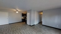 This single-floor space features an open-concept design that seamlessly connects the living area and kitchen. The flooring throughout consists of durable, wood-look planks in a cool grey-brown tone, providing a modern and cohesive aesthetic across the space. The living area is equipped with a ceiling fan, offering both lighting and air circulation. Neutral grey walls create a clean, versatile backdrop throughout.

The kitchen is positioned at the far end of the open layout and is outfitted with dark espresso-toned cabinetry, a black electric range, and a range hood. Light-toned countertops provide contrast against the dark lower and upper cabinets. A partial dividing wall subtly separates the kitchen from the main living space while maintaining an open feel.

A set of sliding closet doors is visible along one wall, offering convenient built-in storage. Adjacent to the closet, a hallway leads further into the unit. The textured popcorn ceiling is consistent throughout, and black baseboards provide a sharp contrast against the light grey walls, adding a contemporary finishing detail to the overall space.