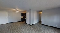 This single-floor space features an open-concept design that seamlessly connects the living area and kitchen. The flooring throughout consists of durable, wood-look planks in a cool grey-brown tone, providing a modern and cohesive aesthetic across the space. The living area is equipped with a ceiling fan, offering both lighting and air circulation. Neutral grey walls create a clean, versatile backdrop throughout.

The kitchen is positioned at the far end of the open layout and is outfitted with dark espresso-toned cabinetry, a black electric range, and a range hood. Light-toned countertops provide contrast against the dark lower and upper cabinets. A partial dividing wall subtly separates the kitchen from the main living space while maintaining an open feel.

A set of sliding closet doors is visible along one wall, offering convenient built-in storage. Adjacent to the closet, a hallway leads further into the unit. The textured popcorn ceiling is consistent throughout, and black baseboards provide a sharp contrast against the light grey walls, adding a contemporary finishing detail to the overall space.