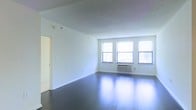 This single-floor space offers a clean, open layout characterized by a minimalist aesthetic and a bright, airy atmosphere. The main living area is anchored by rich, dark hardwood floors that reflect natural light beautifully, creating a polished and inviting ambiance throughout. Three large windows line the exterior wall, flooding the interior with generous amounts of daylight and providing views of the surrounding environment. White walls and white trim throughout the space contribute to a crisp, neutral palette that makes the area feel open and versatile. A textured ceiling adds subtle architectural character to the room. A wall-mounted HVAC unit is conveniently positioned beneath the windows, ensuring year-round climate control. The open plan of the main living area allows for flexible furniture arrangements and a variety of uses. A doorway visible along the interior wall provides access to additional areas of the unit, suggesting a practical and functional layout. The overall atmosphere is bright, clean, and move-in ready, offering a solid foundation for a variety of interior design styles. The space is well-suited for those seeking a straightforward, light-filled environment with classic finishes.