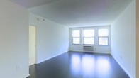 This single-floor space offers a clean, open layout characterized by a minimalist aesthetic and a bright, airy atmosphere. The main living area is anchored by rich, dark hardwood floors that reflect natural light beautifully, creating a polished and inviting ambiance throughout. Three large windows line the exterior wall, flooding the interior with generous amounts of daylight and providing views of the surrounding environment. White walls and white trim throughout the space contribute to a crisp, neutral palette that makes the area feel open and versatile. A textured ceiling adds subtle architectural character to the room. A wall-mounted HVAC unit is conveniently positioned beneath the windows, ensuring year-round climate control. The open plan of the main living area allows for flexible furniture arrangements and a variety of uses. A doorway visible along the interior wall provides access to additional areas of the unit, suggesting a practical and functional layout. The overall atmosphere is bright, clean, and move-in ready, offering a solid foundation for a variety of interior design styles. The space is well-suited for those seeking a straightforward, light-filled environment with classic finishes.