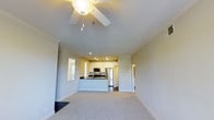 This single-floor space showcases an open-concept design that connects the living area and kitchen in a bright, cohesive layout. The living area features plush carpeting, crown molding, and a vaulted ceiling fitted with a ceiling fan, adding both airflow and visual height to the room. Natural light filters in through a window dressed with blinds, keeping the space feeling airy and well-lit.

The kitchen is positioned at the heart of the unit, defined by a peninsula-style counter with a dark countertop that provides a striking contrast to the white cabinetry above and below. Stainless steel appliances, including a refrigerator, microwave, and range, lend a sleek, modern feel. Recessed lighting illuminates the kitchen area, complementing the clean white cabinetry and neutral color palette.

A hallway visible beyond the kitchen suggests additional rooms, offering functional separation between living and private spaces. Throughout, the neutral tones of the walls and flooring create a versatile, move-in-ready aesthetic. The overall ambiance is clean, modern, and welcoming, well-suited for comfortable everyday living.