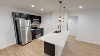 This thoughtfully designed single-floor unit showcases a cohesive modern aesthetic with light hardwood flooring and crisp white walls running throughout. The open-concept layout anchors around a striking kitchen featuring dark cabinetry, a tile backsplash, and a full suite of stainless steel appliances including a refrigerator, electric range, and built-in microwave. A sleek white waterfall-edge kitchen island with an integrated black matte sink serves as the centerpiece, illuminated by a pair of stylish pendant lights that add warmth and visual interest to the space. The open living area extends generously from the kitchen, offering a bright and versatile space ideal for a variety of configurations. Recessed lighting throughout provides consistent, even illumination across all rooms. The bedrooms are well-proportioned with large windows fitted with horizontal blinds that allow for natural light while maintaining privacy. The bathroom features large-format white wall tiles, a full soaking tub with an overhead shower, matte black fixtures, and hexagonal mosaic floor tiles, delivering a polished, on-trend finish. The overall ambiance of the unit is clean, contemporary, and move-in ready, with a neutral palette that provides an excellent canvas for personalization.