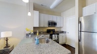 This single-floor residence showcases an open-concept layout that connects the kitchen and living area into one cohesive, functional space. The kitchen is a standout feature, equipped with granite countertops, white cabinetry, and stainless steel appliances, including a refrigerator, range, and built-in microwave. A peninsula with an undermount sink and additional counter space extends into the room, providing both a prep area and a casual gathering spot. Pendant lighting hangs above the peninsula, adding focused illumination and a stylish touch to the space.

The cabinetry is finished in crisp white, offering ample storage, while the dark hardwood flooring throughout creates a warm contrast against the light walls and countertops. The kitchen transitions naturally into the living area, which benefits from natural light streaming through a window. A ceiling fan provides comfort overhead. The overall palette of soft neutrals, complemented by the rich flooring, gives the space a clean, contemporary feel that is both welcoming and practical.
