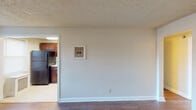 This single-floor unit offers a thoughtfully laid-out living space with a warm, functional design throughout. The living area features rich hardwood flooring that extends across the main rooms, complemented by neutral-toned walls and classic textured ceilings. Multiple windows with blinds allow natural light to filter in, providing pleasant views of the surrounding greenery. A radiator heating system is present in several rooms, adding a classic architectural touch.

The kitchen is a standout feature, showcasing rich dark wood cabinetry with modern hardware, light granite-style countertops, and black stainless steel appliances including a refrigerator and a gas range. A double-basin sink and ample upper and lower cabinet storage complete the space, which is finished with light tile flooring for easy maintenance.

The bedroom offers a comfortable layout with hardwood flooring, a window with outdoor views, and a radiator unit. A separate room provides two closet doors, offering practical storage options. The bathroom is clean and functional, featuring a full-size soaking tub with an integrated white surround and a showerhead. Clean white finishes and neutral walls keep the space feeling fresh and understated throughout.