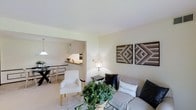 This single-floor residence offers a thoughtfully arranged open-concept layout that seamlessly connects the main living areas. The living room serves as the heart of the home, featuring light neutral walls that create a bright and airy atmosphere. Decorative wooden geometric wall art adds a warm, textural accent to the space, while a glass-top coffee table and a mix of seating — including a plush sofa and a clean-lined white accent chair — provide both comfort and style. A potted indoor plant introduces a touch of greenery, enhancing the welcoming ambiance.

The living area flows naturally into the dining space, which is defined by a pendant light overhead and a glass-top dining table with coordinating chairs, making it an inviting spot for everyday meals. Adjacent to the dining area, the kitchen features a raised breakfast bar countertop in warm wood tones, providing a casual seating or serving area. The kitchen is compact yet functional, with built-in appliances visible along the wall. Throughout the space, light-toned flooring unifies the rooms and contributes to the overall sense of openness and cohesion. The neutral color palette and curated furnishings give the space a modern yet approachable character.